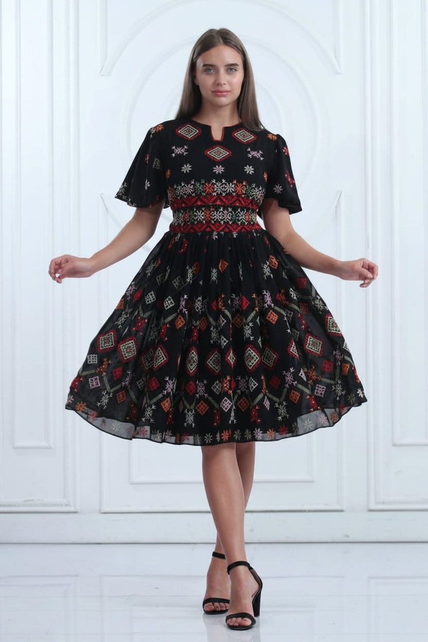 A woman modeling a black short-sleeve A-line dress with colorful geometric patterns and a smocked bodice, standing against a white paneled wall. Arabic text and a circular inset showing the pattern details are visible in the top left corner.