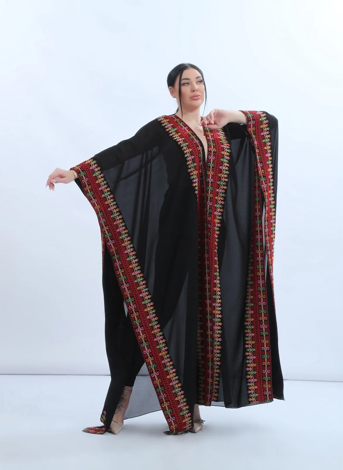 Side drape of the embroidered chiffon bisht showing the flow of the wide open sleeve and the red Tatreez-inspired border, انسدال جانبي للبشت المطرز من الشيفون يظهر حركة الكم الواسع وحدود التطريز الحمراء المستوحاة من التطريز الشامي